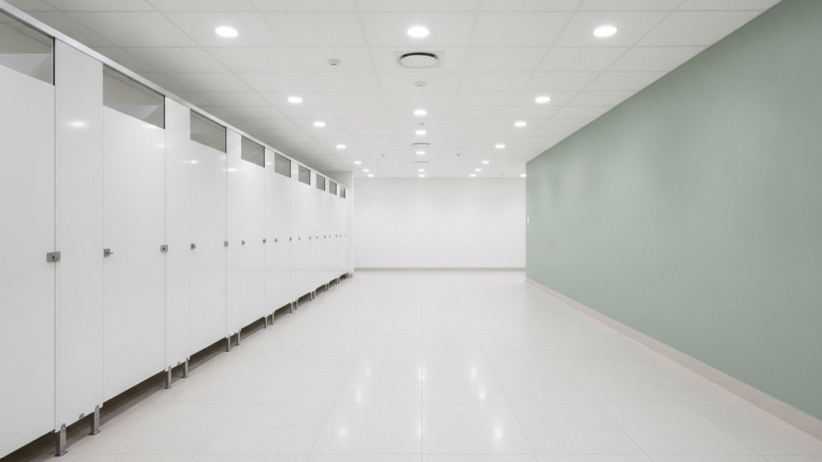 A large corporate office washroom with a long row of white cubicle doors and a sage-green feature wall
